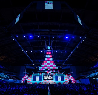 An expansive venue filled with a large audience, featuring a grand stage lit with vibrant blue and pink lights. The ceiling is decorated with a large display, and numerous screens showcase the event's branding. The atmosphere is dynamic and futuristic, reflecting a technologically advanced conference.